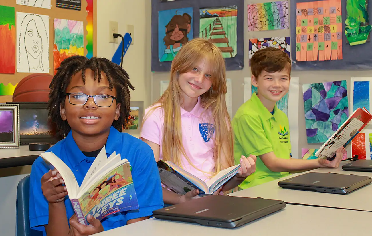 Photo of 3 kids at a desk smiling and reading books.