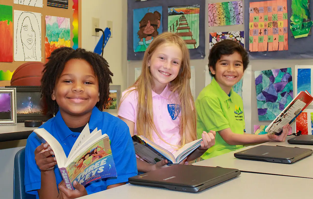 Photo of 3 kids at a desk smiling and reading books.