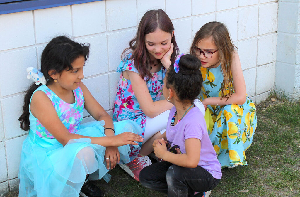 Three 4th grade girls in dresses crouched down talking to a kindergartner